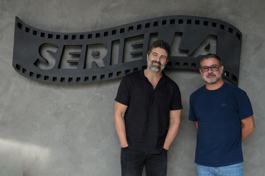 Carlos Manga Jr. ao lado de um homem de camisa azul em frente o logo da Seriella