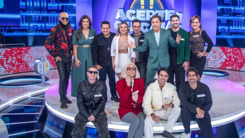 Tom Cavalcante com um terno verde musgo posa com os 11 participantes do reality show "Acerte ou Caia!" no palco, todos sorrindo e em clima de festa.