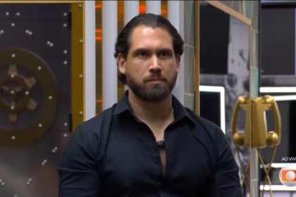 Brigido de camisa preta e semblante assustado ao descobrir que estava eliminado do BBB 26. Ele está sentado no sofá da sala
