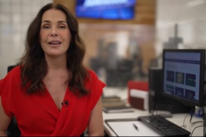 Carolina Ferraz no cenário da redação do Domingo Espetacular. Ela está sentada com vestido vermelho em frente a um computador apresentando os destaques do Domingo Espetacular