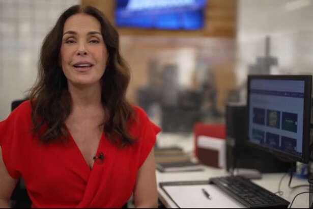 Carolina Ferraz no cenário da redação do Domingo Espetacular. Ela está sentada com vestido vermelho em frente a um computador apresentando os destaques do Domingo Espetacular