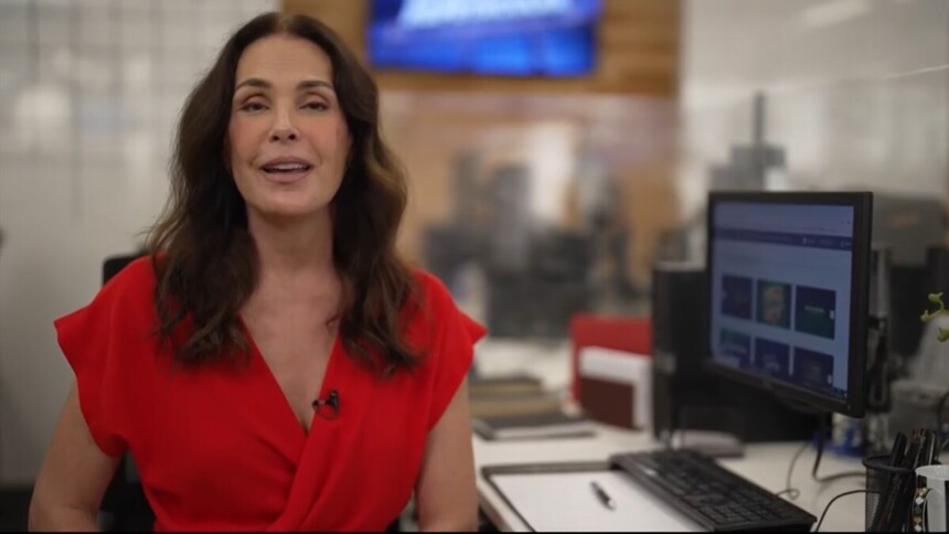 Carolina Ferraz no cenário da redação do Domingo Espetacular. Ela está sentada com vestido vermelho em frente a um computador apresentando os destaques do Domingo Espetacular