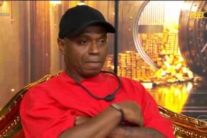 Edilson Capetinha de roupa vermelha e boné preto sentado na cadeira do confessionário esculta a voz da produção anunciando sua expulsão do BBB 26