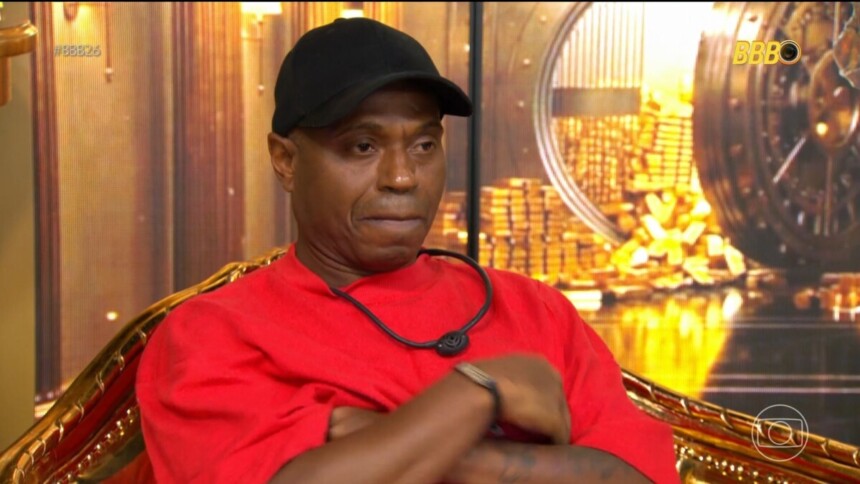 Edilson Capetinha de roupa vermelha e boné preto sentado na cadeira do confessionário esculta a voz da produção anunciando sua expulsão do BBB 26