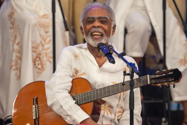 Gilberto Gil sorridente com um violão no colo no palco do Altas Horas