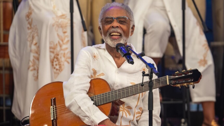 Gilberto Gil sorridente com um violão no colo no palco do Altas Horas