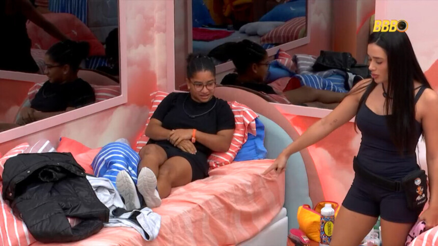 Milena deitada na cama de Jordana usando tênis enquanto a rival observa de pé e as duas discutem no quarto do BBB 26