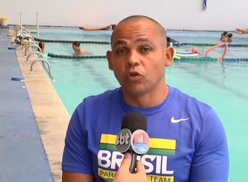 Adriano Gomes Lima sentado em uma cadeira de rodas em frente a uma piscina em entrevista ao microfone do SBT