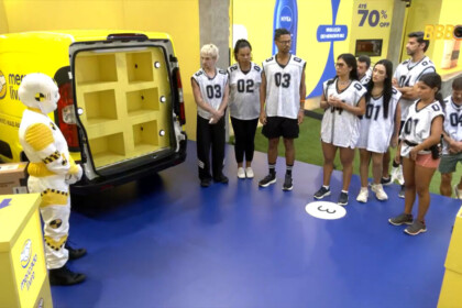 Dummie se posiciona ao lado de uma van no cenário da Prova do Anjo enquanto os participantes observam as instruções no BBB 26