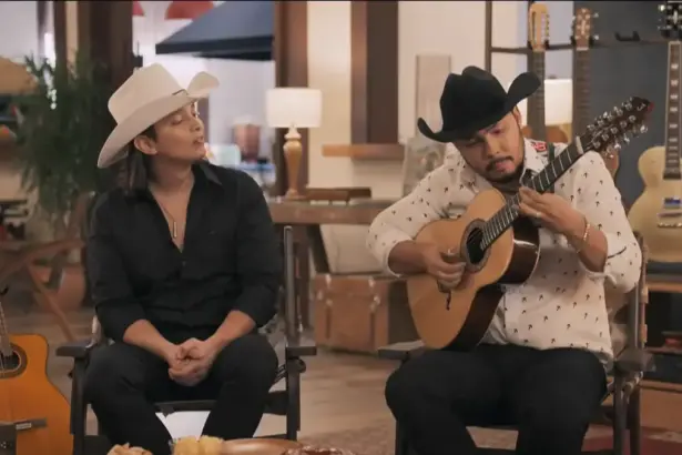 Homem toca violão enquanto outro convidado do Viver Sertanejo observa. Eles estão sentados em um estúdio da fazenda de Daniel