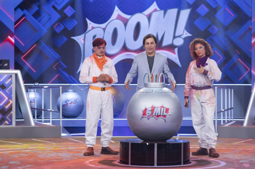 Dudu Camargo, Tom Cavalcante e Bárbara Borges olham assustados para uma bomba prateada no centro do palco do Boom!