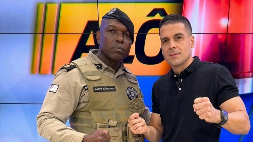 Marcelo Castro, o Juca, segura medalha ao lado do PM no estúdio do programa Alô Juca da TV Aratu, afiliada do SBT. Ambos estão com semblante sério