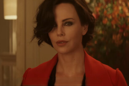Charlize Theron vestida com um sobretudo vermelho, de cabelo curto e com uma arma em mãos em cena do filme Atômica