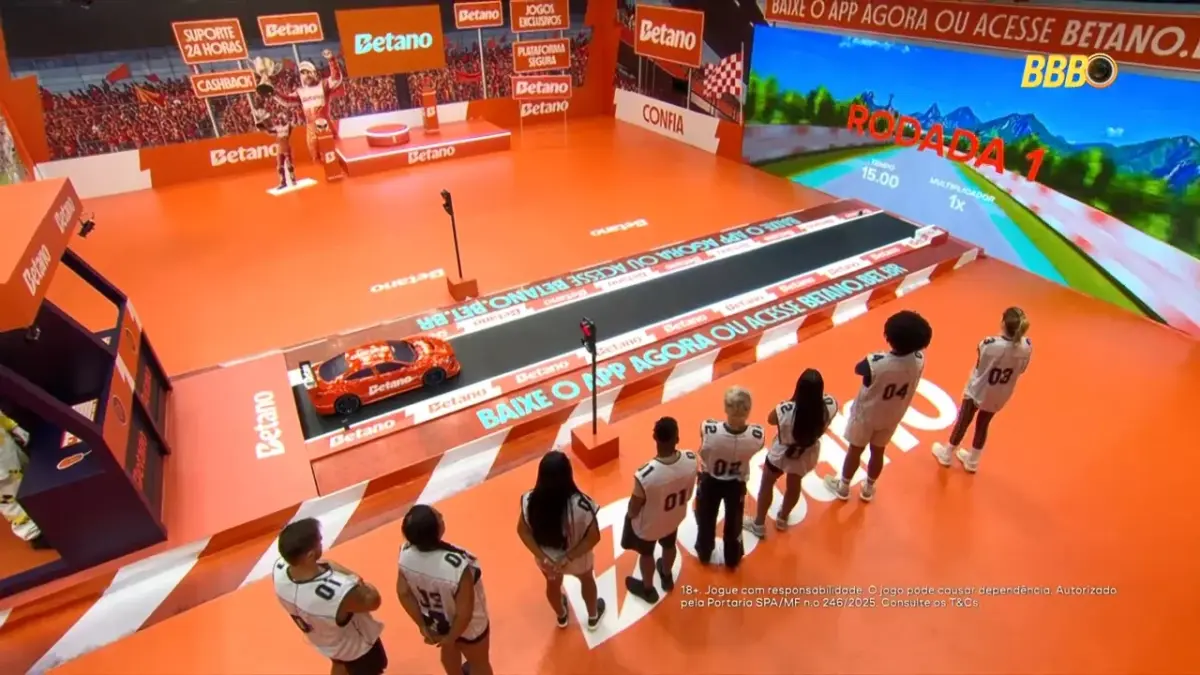 Cenário laranja da Prova do Anjo com um mini carro de corrida no centro e um grande telão para os jogadores