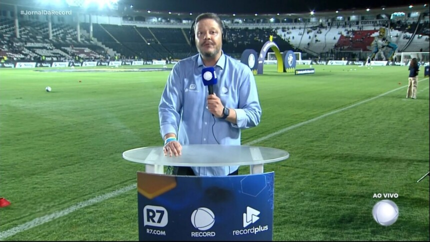 Bruno Laurence segura microfone da RECORD na bancada do Brasileirão montada no meio do campo de futebol