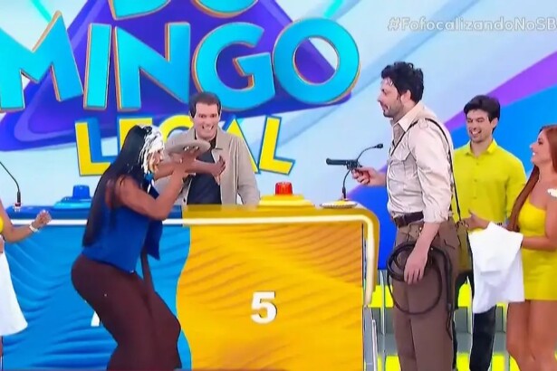 Gracyanne Barbosa se abaixa enquanto Danilo Gentili aponta uma arma para ela no cenário do Domingo Legal do SBT