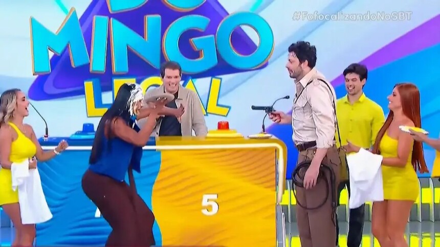 Gracyanne Barbosa se abaixa enquanto Danilo Gentili aponta uma arma para ela no cenário do Domingo Legal do SBT