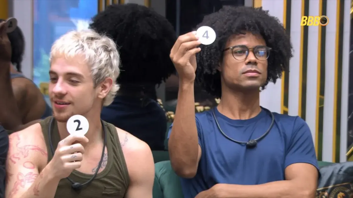 Juliano e Breno seguram números do sorteio da Prova do Anjo. Eles estão sentados na sala do BBB 26