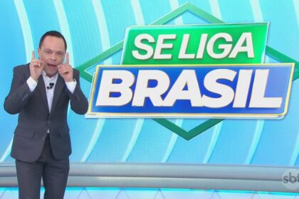 Thiago Gardinali aponta os dois dedos para cima no cenário do Se Liga Brasil no SBT comemorando a alta audiência. Ele está sorridente