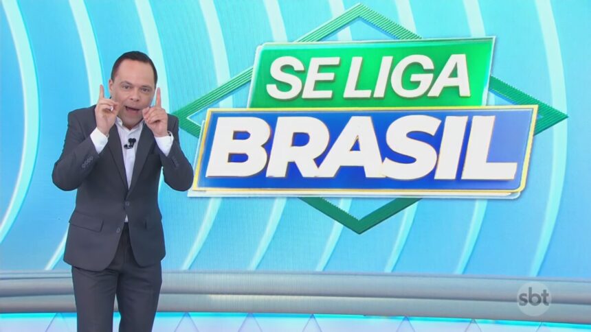Thiago Gardinali aponta os dois dedos para cima no cenário do Se Liga Brasil no SBT comemorando a alta audiência. Ele está sorridente