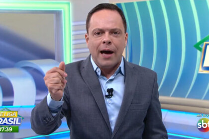 Thiago Gardinali com a boca aberta apresentando o Se Liga Brasil de terno e gravata no cenário que se parece com o Balanço Geral da RECORD