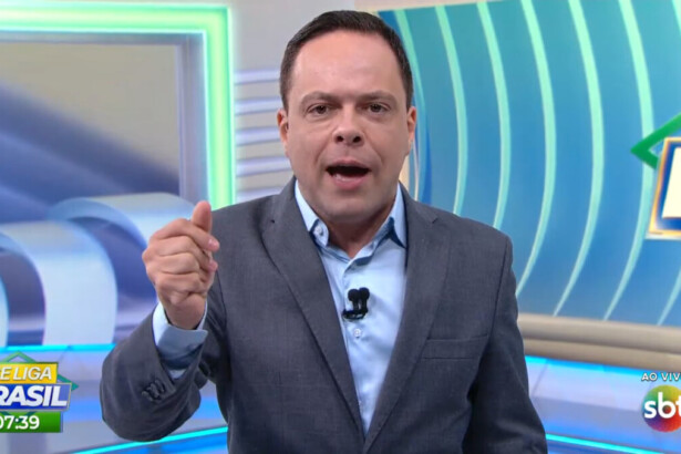 Thiago Gardinali com a boca aberta apresentando o Se Liga Brasil de terno e gravata no cenário que se parece com o Balanço Geral da RECORD