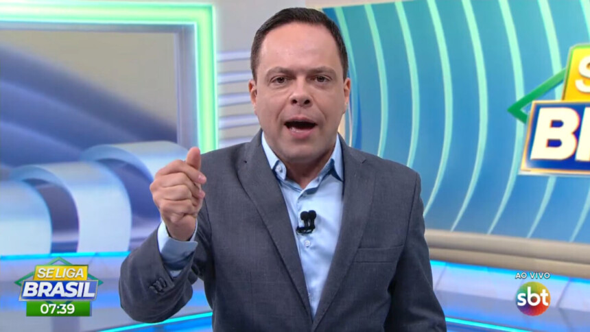 Thiago Gardinali com a boca aberta apresentando o Se Liga Brasil de terno e gravata no cenário que se parece com o Balanço Geral da RECORD
