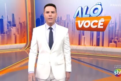 Luiz Bacci veste terno branco e com semblante assustado no cenário do Alô Você