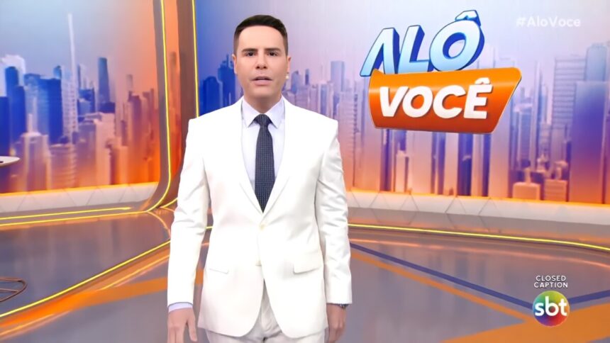 Luiz Bacci veste terno branco e com semblante assustado no cenário do Alô Você