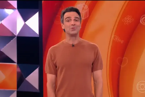 Tadeu vest camisa laranja sorridente no centro do cenário do BBB 26 apresentando a dinâmica da semana