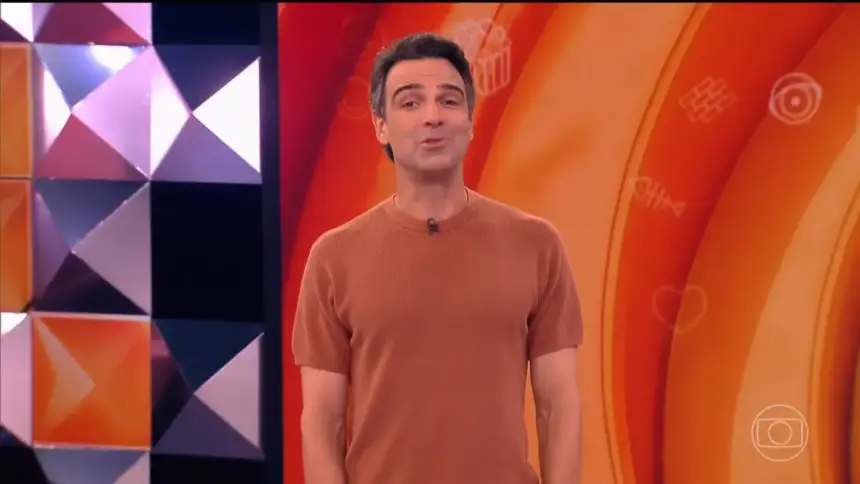 Tadeu vest camisa laranja sorridente no centro do cenário do BBB 26 apresentando a dinâmica da semana