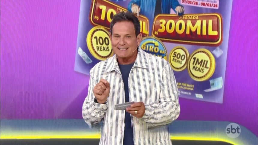 Luís Ricardo no centro do palco da Tele Sena Semanal edição 94. Ele está em frente ao telão que mostra o bilhete lilás