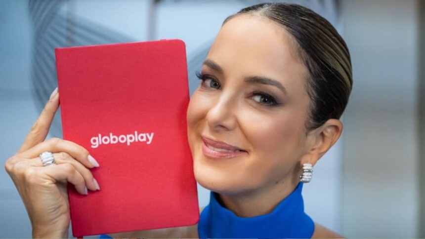 Ticiane Pinheiro sorrindo, segurando próximo ao rosto uma agenda vermelha com o logo do Globoplay