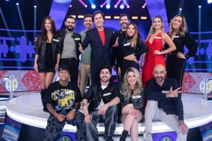 Tom Cavalcante com um terno azul escuro posa com os 11 participantes do reality show "Acerte ou Caia!" no palco, todos sorrindo e em clima de festa.