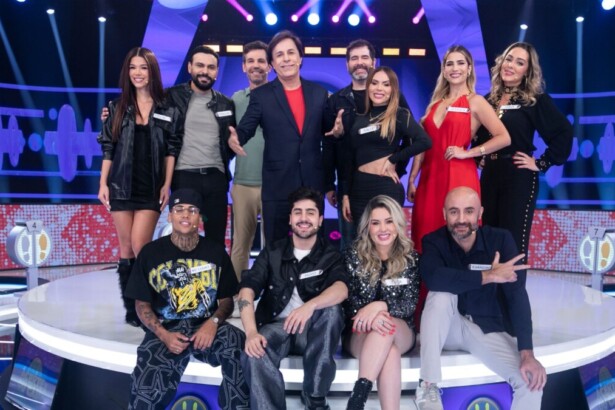 Tom Cavalcante com um terno azul escuro posa com os 11 participantes do reality show "Acerte ou Caia!" no palco, todos sorrindo e em clima de festa.