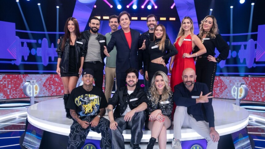 Tom Cavalcante com um terno azul escuro posa com os 11 participantes do reality show "Acerte ou Caia!" no palco, todos sorrindo e em clima de festa.