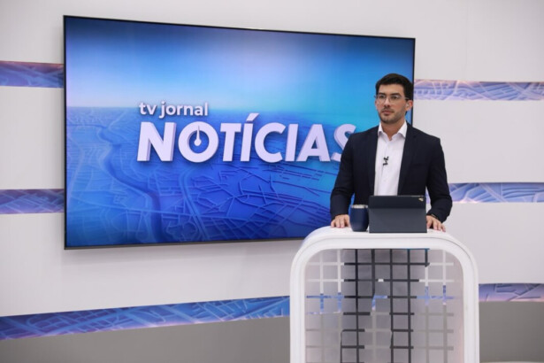 Victor Tavares em frente ao telão com o logo do TV Jornal Notícias e na bancada do telejornal da afiliada do SBT