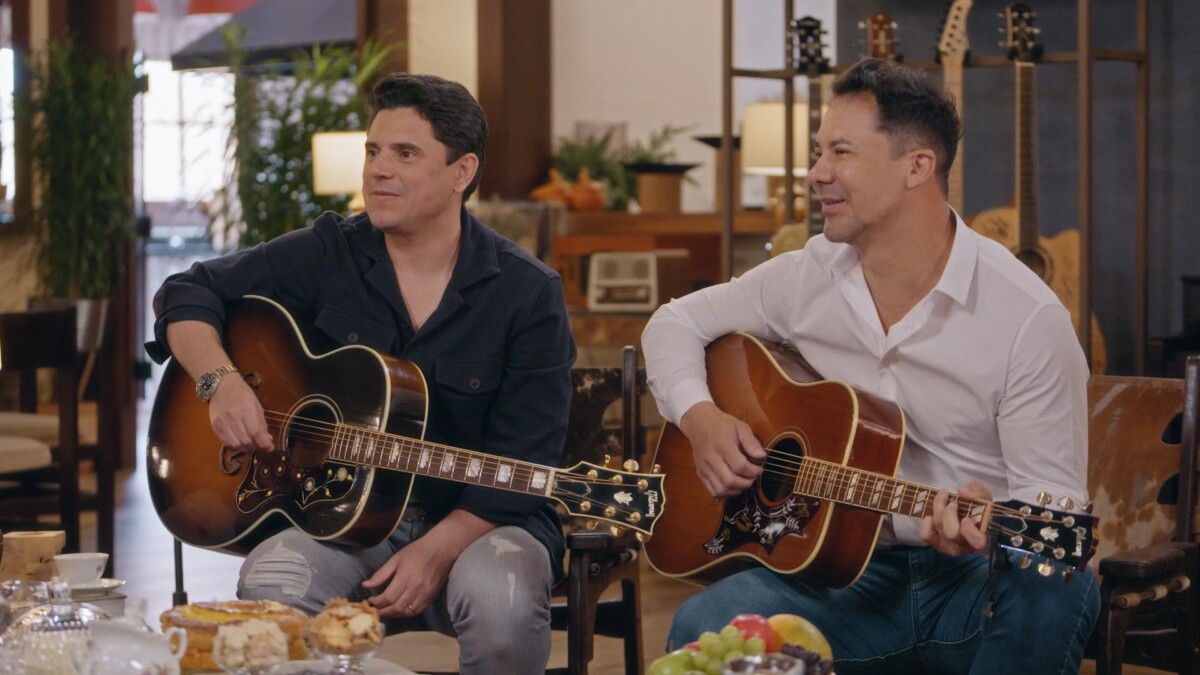 Hugo Pena & Gabriel seguram violões e sorriem enquanto conversa com Daniel