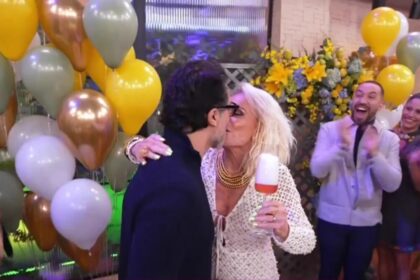 Fábio Arruda de terno escuro beija Ana Maria Braga na boca. Ela está com um vestido branco segurando uma taça no cenário do Mais Você rodeado de amigos e famosos