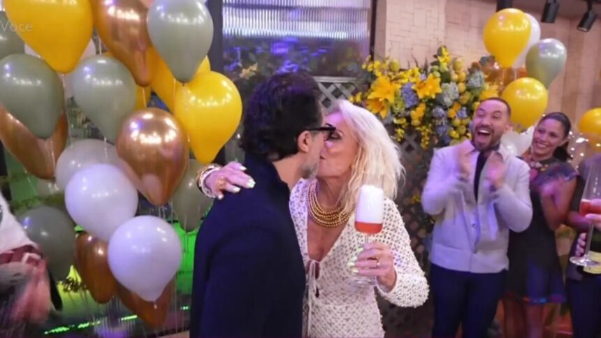 Fábio Arruda de terno escuro beija Ana Maria Braga na boca. Ela está com um vestido branco segurando uma taça no cenário do Mais Você rodeado de amigos e famosos