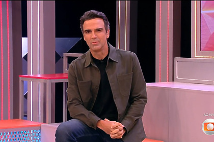Tadeu Schmidt sentado nos blocos do cenário do programa anunciando as incrições do BBB 27