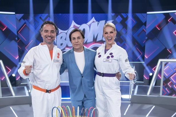 Marco Luque com figurino laranja e Ana Hickman com o roxo abraçam Tom Cavalcante em frente a uma bomba dourada no palco do Boom!