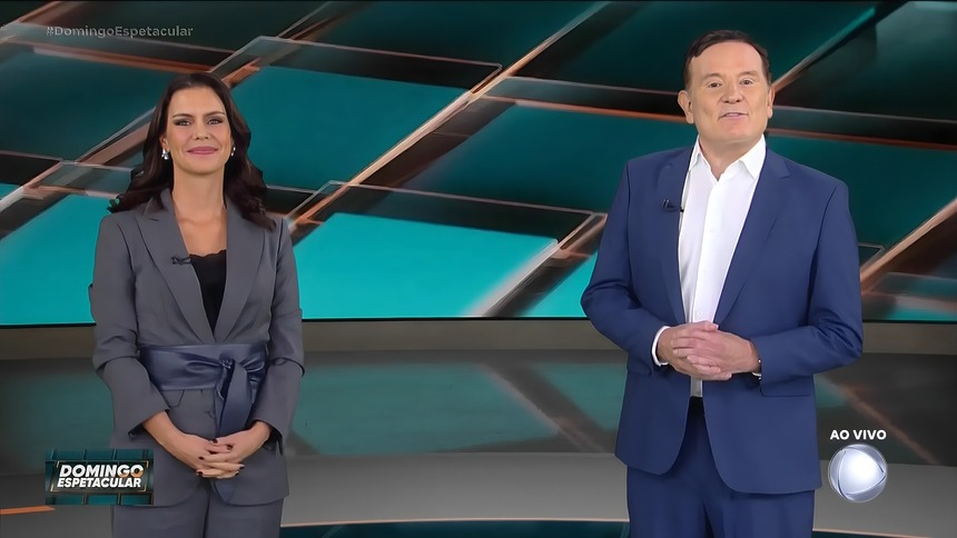 Camila Busnello ao lado de Cabrini. Os dois sorridentes em pé no cenário do Domingo Espetacular