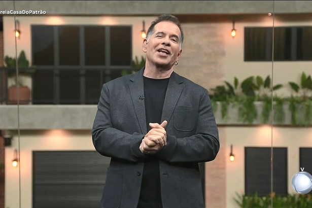 Leandro Hassum com as mãos unidas e sorridente no centro do cenário estúdio glass. Ao fundo a Casa do Patrão