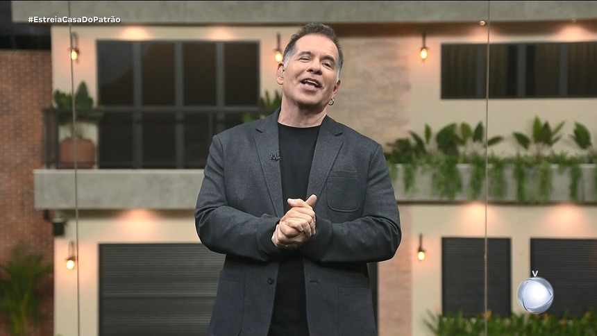Leandro Hassum com as mãos unidas e sorridente no centro do cenário estúdio glass. Ao fundo a Casa do Patrão