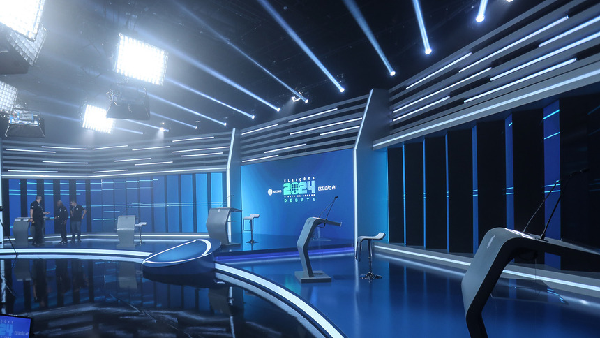 Cenário do Debate das Eleições na RECORD em parceria com o Estadão. Luzes azuis e bancadas em tons de cinza compõe o espaço bem alto e moderno com painéis de LED