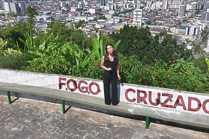 Camila Busnello em cima de um morro, com uma frase digitalizada escrito Fogo Cruzado no muro em chamada do Domingo Espetacular