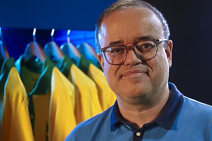 Everaldo Marques com uniforme do esporte da Globo em frente a um vestuário com camisas da seleção brasileira na Copa do Mundo