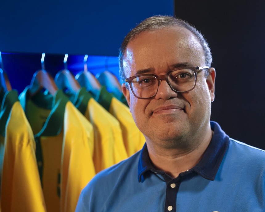Everaldo Marques com uniforme do esporte da Globo em frente a um vestuário com camisas da seleção brasileira na Copa do Mundo