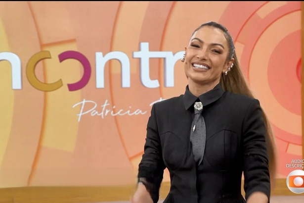 Patricia Poeta com conjunto preto, sorridente, em frente ao telão do programa Encontro